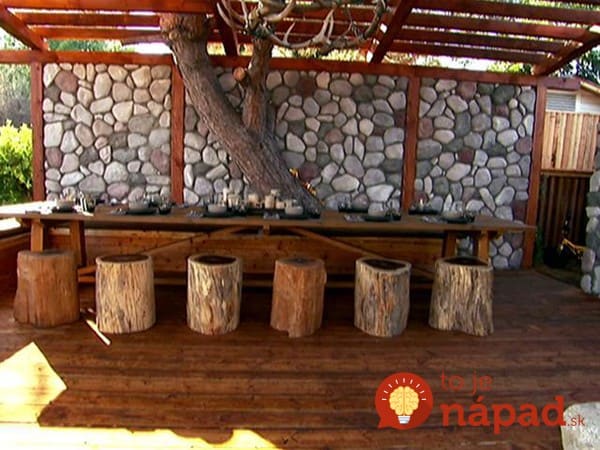 An outdoor banquet table with stools made from trees to look like stumps. Behind the table is a cobblestone wall and wooden posts that hold up the structure above.
